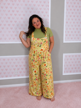 Woman wearing a yellow floral jumpsuit in a room with heart-patterned wallpaper.