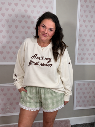 A woman wearing a cream sweatshirt with brown script text reading 'Ain't my first rodeo' and green plaid shorts, standing in front of a wall with pink heart patterns.