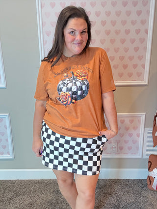 Person wearing an orange fall graphic t-shirt and a checkered skirt in a room with heart-patterned wallpaper.