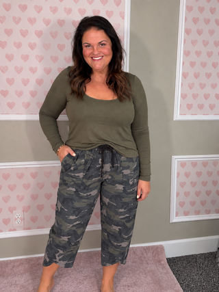A woman modeling a two-piece loungewear set consisting of an olive green long-sleeve top and dark green camouflage cropped pants, standing in a room with pink heart-patterned wallpaper.