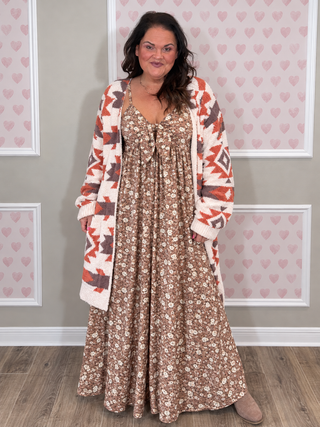 A woman modeling a long-sleeve maxi dress with a brown and beige floral pattern, layered under a cream knit cardigan featuring orange, brown, and black geometric patterns, set against a pink heart-patterned wall.