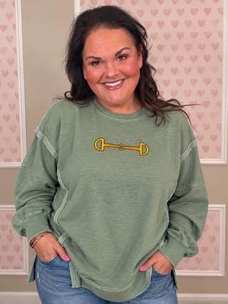 A woman wearing a sage green long-sleeve sweatshirt with a yellow horse bit graphic on the chest, paired with blue jeans, standing in front of a pink heart-patterned wall.