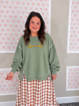 A woman wearing an oversized sage green sweatshirt with a yellow horse bit graphic on the chest, paired with a brown and white gingham skirt, standing in front of a wall with pink heart wallpaper.