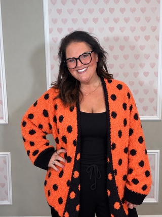 A woman wearing a bright orange fuzzy cardigan with black polka dots over a black top, standing in front of a wall with pink heart wallpaper.