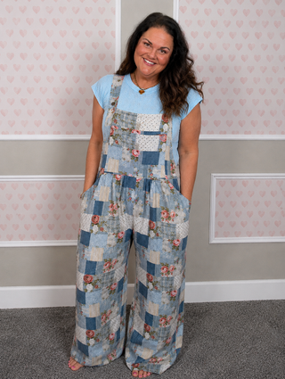 A woman modeling a pair of patchwork blue floral wide-leg overalls over a light blue t-shirt, standing in front of a pink heart-patterned wall.