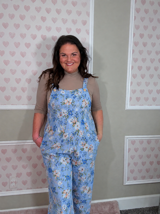 Woman wearing a blue floral jumpsuit standing in a room with heart-patterned wallpaper.