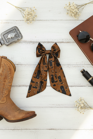 Brown satin bow with pattern, cowboy boot, notebook, and pen on a white wooden surface.