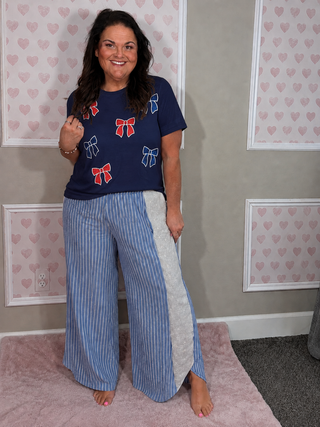 A woman modeling a navy blue t-shirt with red and blue bow prints and matching blue-and-white striped wide-leg pants with a gray side panel, standing in a room with pink heart-patterned wall panels.