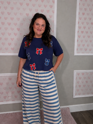 A woman modeling a navy blue short-sleeve t-shirt with red and blue bow prints, paired with blue and white striped pants, standing in front of a gray wall with pink heart wallpaper.