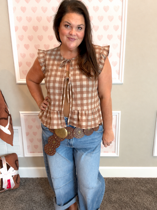 A woman modeling a sleeveless brown and beige gingham tie-front top with ruffled shoulders, paired with light blue jeans and a decorative brown belt, standing in front of a wall with framed pink heart artwork.
