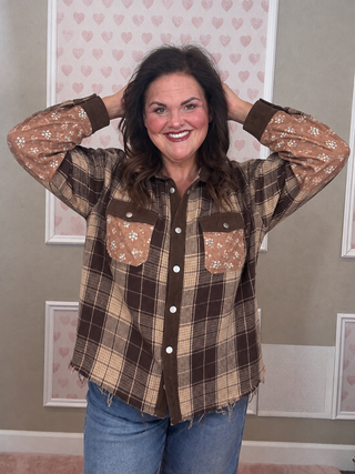 A woman modeling a brown and beige plaid button-up shirt with floral-patterned pockets and sleeves, paired with blue jeans, standing against a decorated wall.
