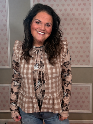 A smiling woman with long dark wavy hair wearing a beige and brown gingham long-sleeve blouse with a horse print overlay, standing in front of a pink heart-patterned wall.