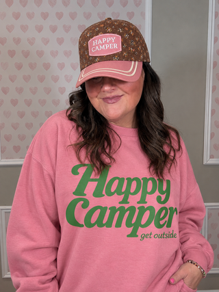 A woman wearing a pink crewneck sweatshirt with green 'Happy Camper' text and a matching brown patterned baseball cap with a pink 'Happy Camper' patch, posed against a heart-patterned wall.