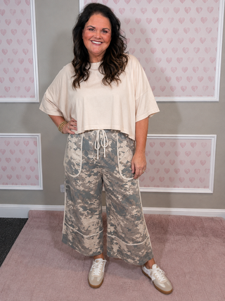 A woman modeling a cream short-sleeve crop top and camouflage wide-leg pants with white sneakers, standing against a pink heart-patterned wall.