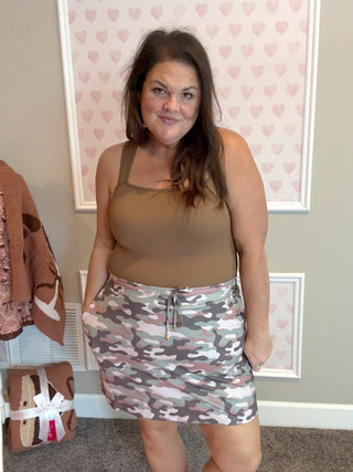 Woman wearing a brown tank top and camouflage skirt indoors with heart-patterned wallpaper.