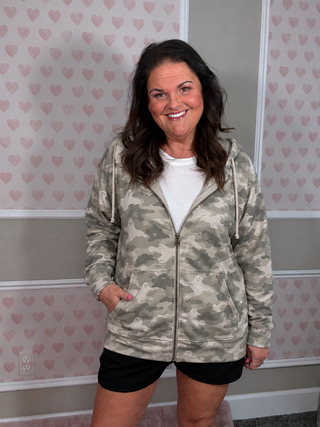A woman wearing a light olive green and gray camouflage zip-up hoodie over a white t-shirt and black shorts, standing in front of a pink heart-patterned wallpaper background.