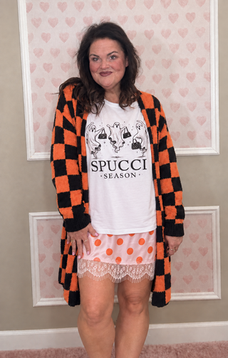 A woman modeling a cozy outfit consisting of a black and orange checkered cardigan, a white graphic t-shirt with 'SPUCCI SEASON' text, and pink polka-dot pajama shorts with lace trim, set against a heart-patterned wall.