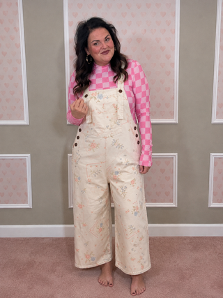A woman stands barefoot indoors wearing cream floral wide-leg overalls over a pink and white checkered long-sleeve top. The room has pink heart-patterned wallpaper and a pink carpet.