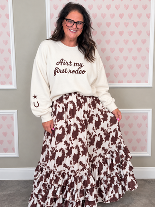 A woman wearing a cream sweatshirt with brown text that reads 'Ain't my first rodeo' and a brown-and-cream cow-print tiered skirt, standing in front of a wall with pink heart patterns.
