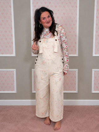 A woman modeling a pair of cream floral print overall pants with a matching floral long-sleeve top, standing in front of a pink and white paneled wall.