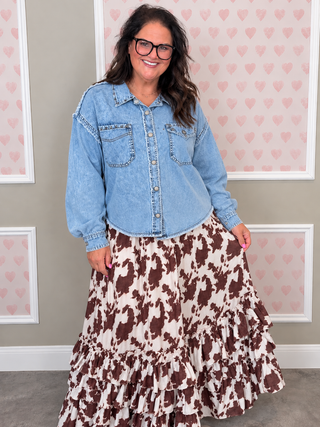 A woman wearing a light blue denim button-up shirt and a brown and cream cow-print ruffled maxi skirt, standing in front of a wall with pink heart wallpaper.