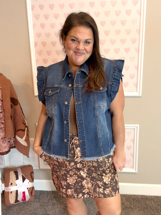 Woman wearing a denim vest over a floral dress in a room with heart-patterned wallpaper.