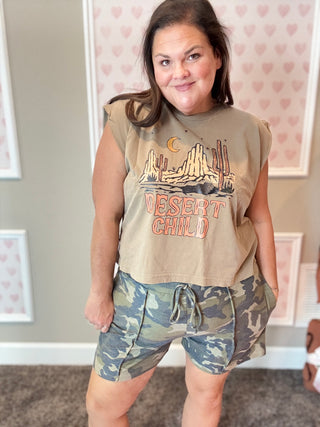 Person wearing a 'Desert Child' t-shirt and camouflage shorts in a room with heart-patterned wallpaper.