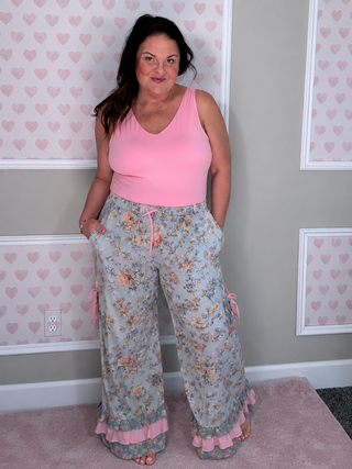 A woman modeling a pink sleeveless top and wide-leg floral pants with pink ruffle hems, standing in front of a gray wall with pink heart-patterned panels.