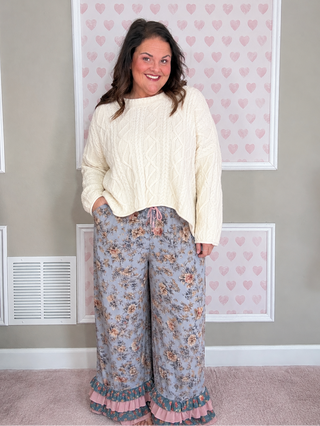 A woman stands indoors wearing a cream cable-knit cropped sweater and wide-leg floral pants with ruffled hems.