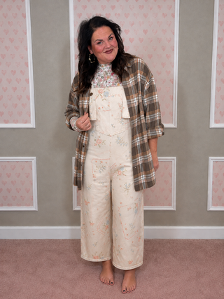 A woman stands indoors wearing a cream floral overall jumpsuit with a matching floral mock neck, a brown and beige plaid flannel shirt, and gold hoop earrings. She is posed against a wall with pink patterned panels and a pink carpet.