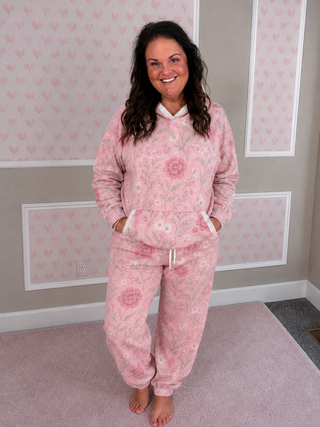 A woman wearing a pink floral fleece pajama set with a hooded sweatshirt and matching jogger pants, standing barefoot in a room with pink heart-patterned wallpaper.