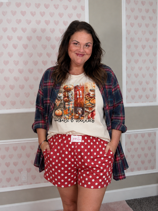 A woman stands indoors wearing a cream graphic T-shirt under a red and navy plaid flannel shirt, paired with red shorts covered in white polka dots. The wall behind her has pale pink heart-patterned wallpaper.