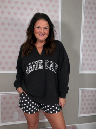 A woman modeling a black long-sleeve V-neck sweatshirt with 'GAME DAY' printed across the front in white block letters, paired with black shorts featuring white polka dots. She is standing in front of a gray wall with decorative panels containing a pink heart pattern.