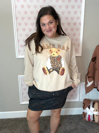 Person wearing a beige sweatshirt with a graphic of a bear and text, standing in a room with heart-patterned wallpaper.
