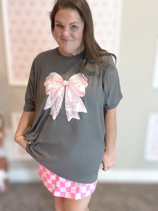Woman wearing a gray t-shirt with a large pink bow and a pink checkered skirt.