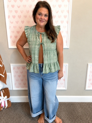 A woman modeling a sage green plaid sleeveless top with a keyhole cutout and ruffle details, paired with light blue jeans, standing in front of a decorated wall with pink heart frames.