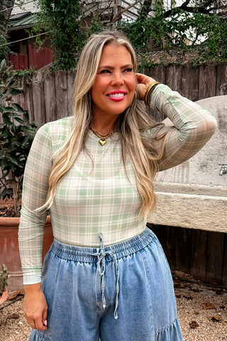 Woman in a plaid shirt and blue jeans standing outdoors with plants and a wooden fence in the background.