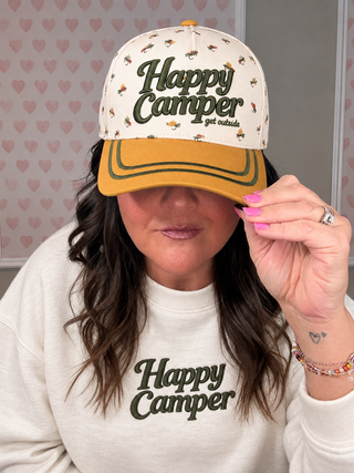 A woman wearing a cream and mustard yellow baseball cap with 'Happy Camper' printed on it, paired with a matching cream sweatshirt. The background features a pink heart patterned wallpaper.