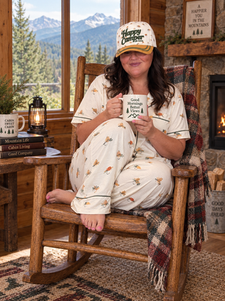 A woman sits in a rustic wooden rocking chair in a cabin, wearing a matching cream pajama set with a small bee print and a 'Happy Camp3r' baseball cap, holding a mug that reads 'Good Mornings Better Sleep'.