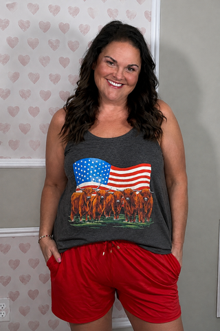 A dark heather gray racerback tank top hanging, featuring a graphic print of Highland cows in front of a waving American flag
