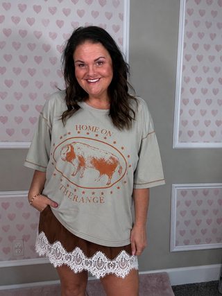 A woman wearing a light sage green graphic t-shirt with a brown cow design and the words 'HOME ON THE RANGE', paired with a brown skirt featuring a white lace hem, standing in front of a wall with pink heart wallpaper.