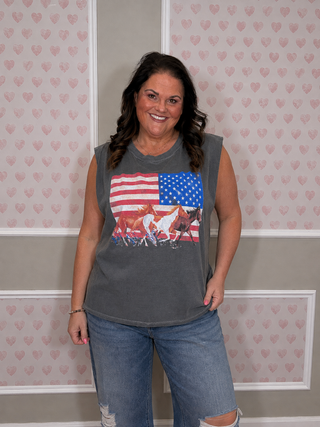 A woman wearing a gray sleeveless graphic t-shirt with a horse and American flag print, paired with distressed blue jeans, standing against a patterned wallpaper background.