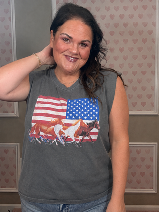 A woman wearing a dark gray sleeveless t-shirt with a graphic of horses in front of an American flag, standing in front of a patterned pink and white heart wallpaper.