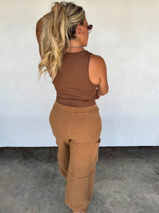 Woman wearing a brown sleeveless top and pants against a white wall.