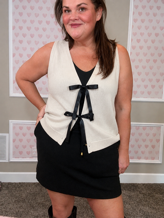 Woman wearing a white sleeveless top with a black bow and black skirt in front of a heart-patterned wall.