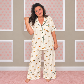 A woman modeling a two-piece pajama set featuring a cream-colored base with a yellow and orange cartoon pattern, accented with dark green piping. She is standing in a room with a pink carpet and a beige wall decorated with pink heart-patterned panels.