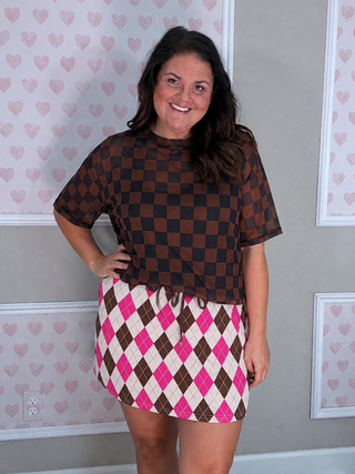 A woman modeling a brown and black checkered short-sleeve top paired with a pink, white, and brown argyle mini skirt, standing in front of a gray wall with pink heart-patterned wallpaper.