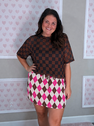 A woman modeling a brown and black checkered short-sleeve top paired with a pink, white, and brown argyle mini skirt, standing in front of a gray wall with pink heart-patterned wallpaper.