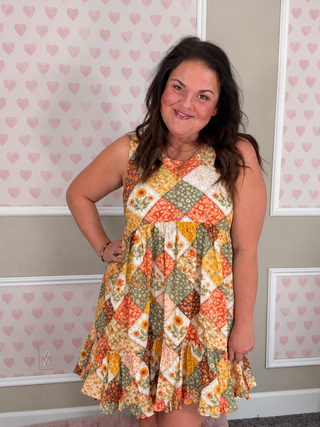 A woman wearing a sleeveless tiered dress with a colorful patchwork print of orange, yellow, green, and white floral and geometric patterns, standing in front of a wall with pink heart wallpaper.