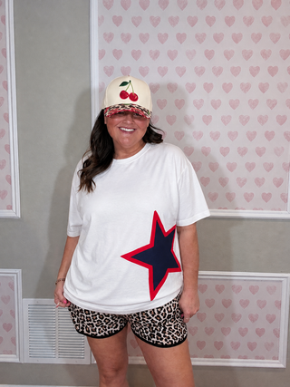 Oversized white patriotic star tee with leopard shorts and cherry trucker hat, playful boutique style outfit

